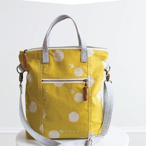 May include: A yellow tote bag with white polka dots and a gray and white shoulder strap. The bag has a zipper closure and a small pocket on the front. The bag is sitting on a white table with the text "noodlehead" written on the table.