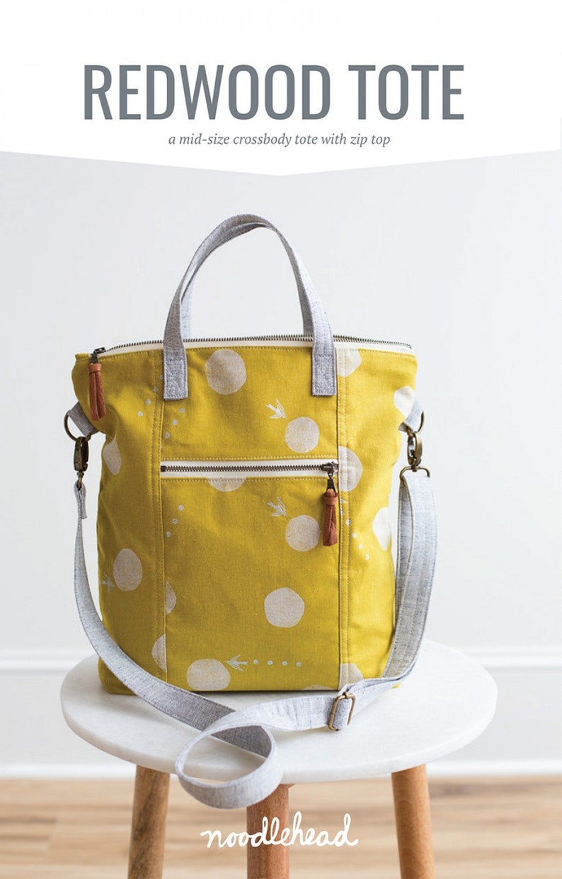 May include: A yellow tote bag with white polka dots and a gray and white shoulder strap. The bag has a zipper closure and a small pocket on the front. The bag is sitting on a white table with the text "noodlehead" written on the table.