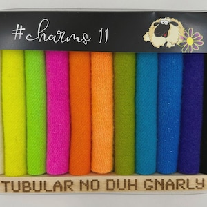 Op de afbeelding: Een set van elf kleurrijke haar scrunchies in een doorzichtige plastic doos. De scrunchies zijn in verschillende kleuren, waaronder wit, geel, groen, roze, oranje, blauw en zwart. De doos heeft de tekst "#charms 11" en "TUBULAR NO DUH GNARLY".