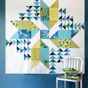 Puede incluir: Un patrón de colcha llamado "Wind Drifter" de Robin Pickens Quilt Patterns. La colcha está hecha con una variedad de telas azules, verdes y amarillas. La colcha se exhibe en una pared azul con una silla blanca y recortes de tela.