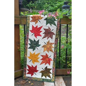 May include: A white table runner with a green, brown, and orange leaf pattern. The runner is decorated with a green, brown, and orange border.