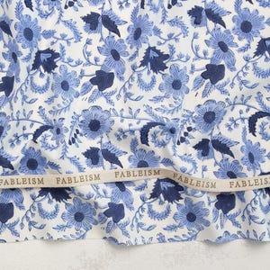 May include: Close-up of a fabric with a floral pattern in shades of blue and white. The design features stylized flowers and leaves. A ribbon with the word "FABLEISM" is visible across the bottom of the fabric.