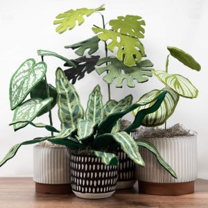 May include: A collection of three potted plants with green felt leaves. The plants are in different styles of pots, including a white pot with a ribbed texture, a black and white patterned pot, and a white pot with a ribbed texture.