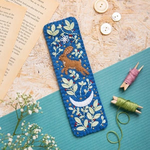 May include: A rectangular blue felt bookmark featuring hand-embroidered details including a brown rabbit, a white crescent moon, and various green leafy branches with small red berries. The edges are finished with a decorative blanket stitch.
