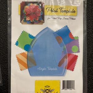 Puede incluir: Una plantilla de acrílico azul para hacer una almohada de zinnia con pétalos pop. La plantilla tiene forma de pétalo y tiene las palabras "Acrylic Template" impresas en ella. La imagen también incluye una pequeña muestra de la almohada terminada, hecha con recortes de tela coloridos.