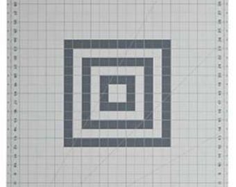 May include: A large, rectangular gray cutting mat with a grid pattern and measurements along the edges. A series of concentric gray squares are centered on the mat. The mat has a hanging hole at the top.