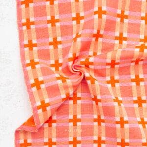 Puede incluir: Un textil plegado de color naranja y rosa con un patrón de rejilla de signos más. La tela tiene un diseño de trama cruzada con diferentes tonos de naranja y rosa. El textil está sobre una superficie blanca.