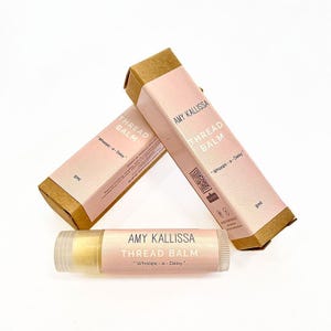May include: Three pink and brown cardboard boxes with the text "AMY KALLISSA THREAD BALM" and "Whoops-a-Daisy" on them. A clear tube of lip balm with the same text is in the foreground.