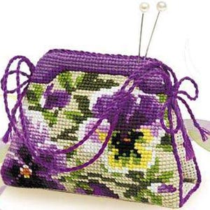 May include: A handmade needlepoint pincushion in a rectangular shape, featuring a floral design with purple, green, white, and yellow hues. The pincushion has a purple border and drawstring closure. Two pearl-headed pins are inserted into the top.