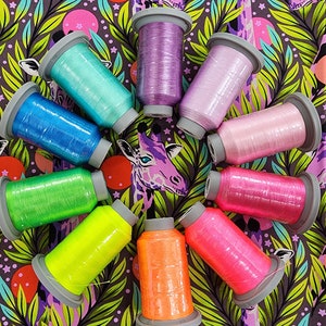 May include: A collection of colorful embroidery thread spools arranged in a circle. The spools are various shades of blue, green, yellow, orange, pink, and purple. The background features a fabric with a giraffe and leaf pattern.