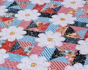 Op de afbeelding: Een quiltpatroon met de titel "blossoms" met een wit madeliefje-ontwerp op een rode, blauwe en zwarte patchwork-achtergrond. Het patroon wordt beschreven als "een 10-vierkant-vriendelijk quiltpatroon van Amanda Murphy Design."