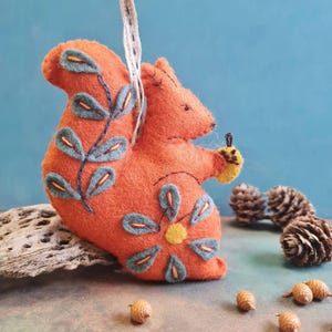 May include: A handmade orange felt squirrel ornament with blue and yellow embroidered details. The squirrel is holding a yellow acorn and has a white ribbon for hanging. Decorative pinecones and acorns are scattered around the ornament.