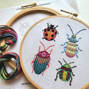 May include: A close-up of a completed embroidery project featuring four colorful embroidered insects on a white fabric stretched within a wooden hoop. The insects include a ladybug, a beetle, and two other stylized bugs. Embroidery floss and a pattern are also visible.