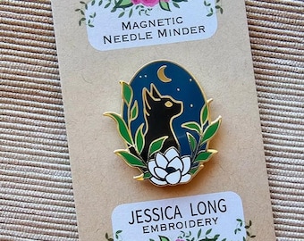 May include: A black cat with yellow eyes looking up at a crescent moon in a blue oval with green leaves and a white flower. The oval is on a brown card with the text "Magnetic Needle Minder" and "Jessica Long Embroidery" with a website address.