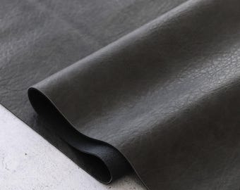 Op de afbeelding: Een close-up van een donkergrijze plaat imitatieleer, met een getextureerd oppervlak en een lichte glans. Het materiaal is opgerold, waardoor de onderkant zichtbaar is. De achtergrond is een lichtgrijs oppervlak.