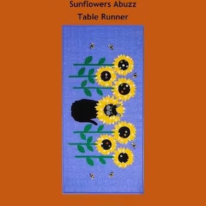 May include: A rectangular table runner with a blue background features a design of yellow sunflowers, green stems, and small black bees. The text "Trouble & Boo Designs", "Sunflowers Abuzz", and "Table Runner" are at the top.