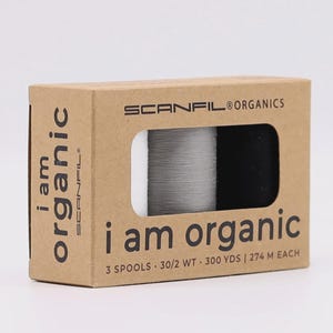 Könnte beinhalten: Eine Pappschachtel mit SCANFIL® ORGANICS Garnspulen. Auf der Schachtel steht der Text "i am organic". Die Schachtel enthält drei Garnspulen mit einer Mischung aus schwarzem und weißem Garn. Jede Spule enthält 274 Meter Garn.
