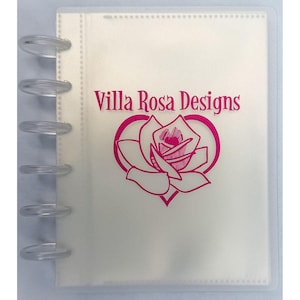 May include: A translucent disc-bound notebook with a frosted cover. The front features the text Villa Rosa Designs in magenta above a magenta line-art illustration of a rose centered within a heart shape. The binding consists of six clear plastic discs.