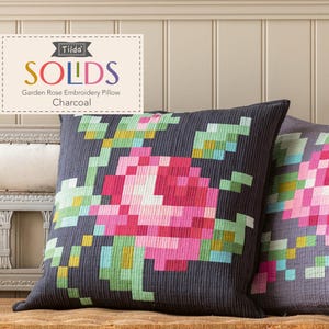 May include: Two square charcoal-colored pillows with a pixelated floral design. The design features a large pink rose with green and yellow leaves. The pillows are labeled "Tilda SOLIDS Garden Rose Embroidery Pillow Charcoal."
