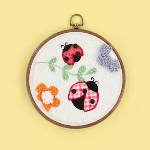 May include: A round, white fabric embroidery hoop featuring a ladybug design. The ladybugs are red and black, with a gingham pattern on one. Other elements include a flower and a blue cloud. The hoop has a wooden frame and a brass hanger.