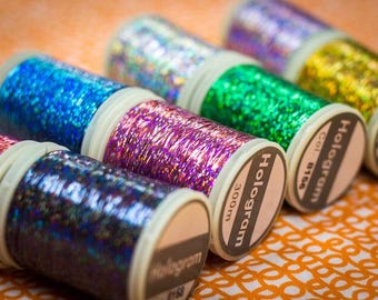 Puede incluir: Primer plano de ocho carretes de hilo holográfico iridiscente. Los carretes están etiquetados como "Hologram" y tienen diferentes colores, incluidos azul, morado, verde y dorado. Cada carrete tiene 300 m de largo.