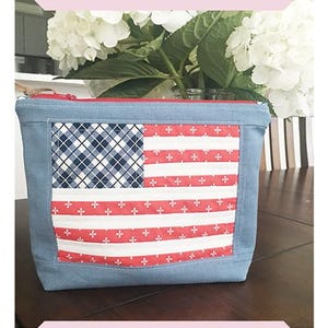 Op de afbeelding: Een blauwe ritszak met een Amerikaans vlagontwerp. De vlag is gemaakt van rood, wit en blauw stof met een geruit patroon in het blauwe veld. De zak is 9 inch bij 10 inch.