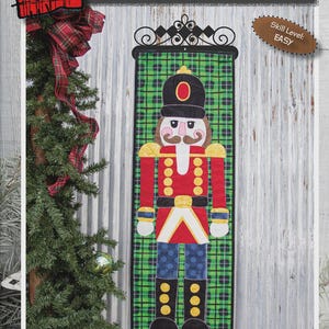 Könnte beinhalten: Ein Wandbehang aus Stoff mit einem Nussknacker-Design. Der Nussknacker trägt eine rote Jacke, blaue Hose und schwarze Stiefel. Der Hintergrund ist grünes Karomuster. Der Text "BIG NUTCRACKER" und die Maße 30.5cm x 84cm sind abgebildet.