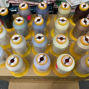 Puede incluir: Una colección de carretes de hilo en varios colores, incluyendo blanco, beige, marrón, azul y gris. Los carretes están dispuestos en filas en un estante de madera. Cada carrete tiene una etiqueta amarilla con la palabra "WonderFil" impresa en ella.