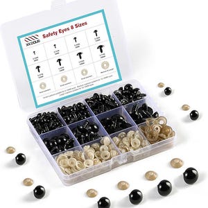 May include: A clear plastic organiser box containing black safety eyes and tan washers in various sizes. The lid shows a chart with eye sizes in millimetres. Several black eyes and tan washers are scattered around the box.