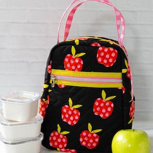 Puede incluir: Una bolsa de almuerzo acolchada negra con estampado de manzanas rojas, asas de cuadros rosas y un bolsillo con cremallera verde lima. El texto "Lunch Box Delight" aparece arriba. Tres recipientes cuadrados blancos, una manzana verde y cubiertos rosas.