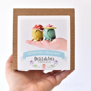 May include: A cardboard box with a white label that says "Stuffed Frog Kit" and "Delilah Iris Designs". The label also features two small felt frogs wearing hats. One frog is green and wearing a pink hat, and the other frog is yellow and wearing a red mushroom hat.