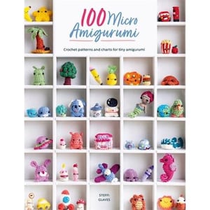 May include: A book titled "100 Micro Amigurumi" with a white background and a colorful display of crocheted amigurumi toys in a white shelf. The book cover features the title in pink and white lettering and the subtitle "Crochet patterns and charts for tiny amigurumi".