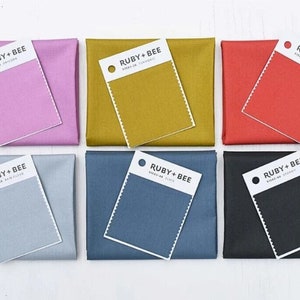 Könnte beinhalten: Sechs Stoffmuster mit Farbkarte, jede mit einer anderen Farbe: rosa, senfgelb, rot, hellblau, dunkelblau und schwarz. Die Karten sind mit "RUBY+BEE" beschriftet, darunter der Text "SHIRTING FABRIC".