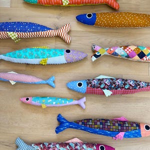 May include: A collection of colorful, patterned fabric fish toys. Each fish features unique designs, including stripes, polka dots, and floral prints, with contrasting fins and eye details. The toys are arranged on a light-colored wooden surface.