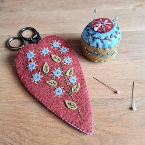 May include: A red felt scissor case, embroidered with blue flowers and green leaves, and a small, round pincushion with blue and brown detailing. Two pins and a pair of scissors are also visible. The items are on a wooden surface.