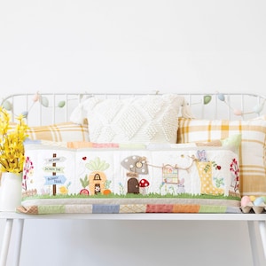 May include: A decorative Easter-themed pillow with a patchwork design. The pillow features embroidered scenes of a bunny trail, a mushroom house, and a bunny in a rain boot. The color palette includes pastels and earth tones.