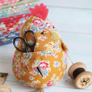 May include: A yellow floral fabric chicken with a red felt comb, black eyes, and a yellow felt beak. The chicken has a pocket on its side with a pair of black scissors sticking out.
