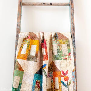 Peut inclure: Une courtepointe en patchwork présentant un motif de maison répétitif dans diverses couleurs, notamment jaune, orange, vert et bleu, sur un fond crème avec des motifs de matelassage floraux. La courtepointe est drapée sur une échelle en bois rustique.
