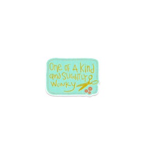 May include: A teal sticker with the text "One of a Kind and Slightly Wonky" in yellow script. The sticker features a pair of gold scissors and three orange and red buttons. The sticker has a white border.