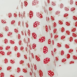 May include: Clear cellophane wrap with a repeating pattern of red mushrooms with white polka dots. The mushroom caps are red with white spots, and the stems are beige. The wrap is slightly translucent.