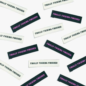 以下が含まれることがあります： ピンクで「FINALLY FUCKING FINISHED」と印刷された黒と白の布製ラベルのコレクションです。