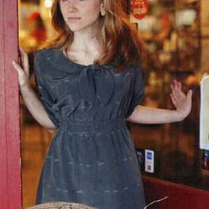 Pode incluir: Um vestido cinza com colarinho branco e laço. O vestido tem cintura franzida e saia fluida. O vestido é etiquetado como "Brynna Dress" e "Sew Liberated" com o número "122".