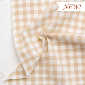 May include: Tan and white gingham fabric with a soft, textured appearance. The fabric is folded, creating shadows and depth. The word "NEW!" is displayed in a red font. The fabric is suitable for various sewing and crafting projects.