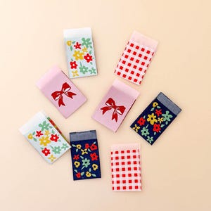 May include: A collection of seven rectangular woven fabric labels featuring various designs including red and white gingham checks, pink backgrounds with red bows, and blue or light blue backgrounds with colorful red, yellow, and green floral patterns.