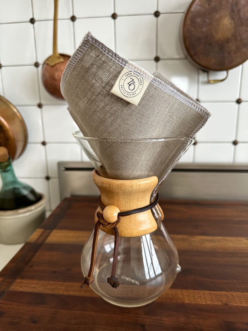 May include: A glass coffee carafe with a wooden collar and a leather strap. A beige linen coffee filter is placed inside the carafe. The carafe is sitting on a wooden surface. The label on the filter reads "The Linen Coffee Filter Co. 710"