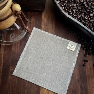 May include: A light gray linen coffee filter with a white stitched border and a small white label with the text "7th & Grove" printed on it.