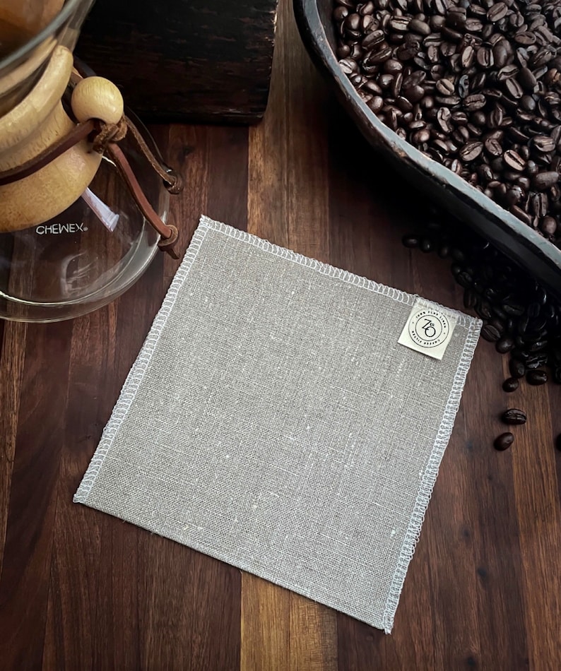 May include: A light gray linen coffee filter with a white stitched border and a small white label with the text "7th & Grove" printed on it.