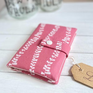 May include: A pink fabric wallet with a white leaf pattern and a white button closure. The wallet is folded closed and has a brown tag with the word "Love" written on it.
