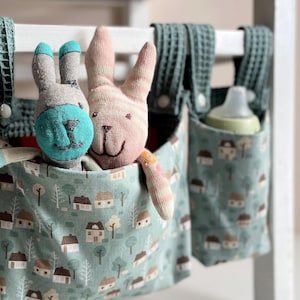 Könnte beinhalten: Zwei hängende Stofftaschen in Salbeigrün mit einem Muster aus kleinen Häusern und Bäumen. Eine Tasche enthält ein graues und türkisfarbenes Socken-Hasenspielzeug und ein rosa gestreiftes Hasenspielzeug. Die andere Tasche enthält eine weiße Babyflasche.
