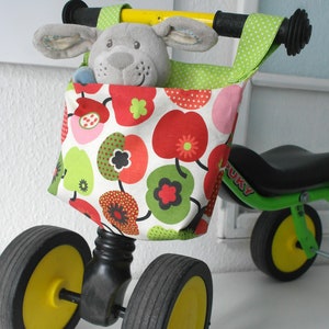 May include: A green and black tricycle with yellow wheels and a fabric storage bag with a red, green and white floral pattern. The bag is attached to the handlebars and contains a grey stuffed animal.